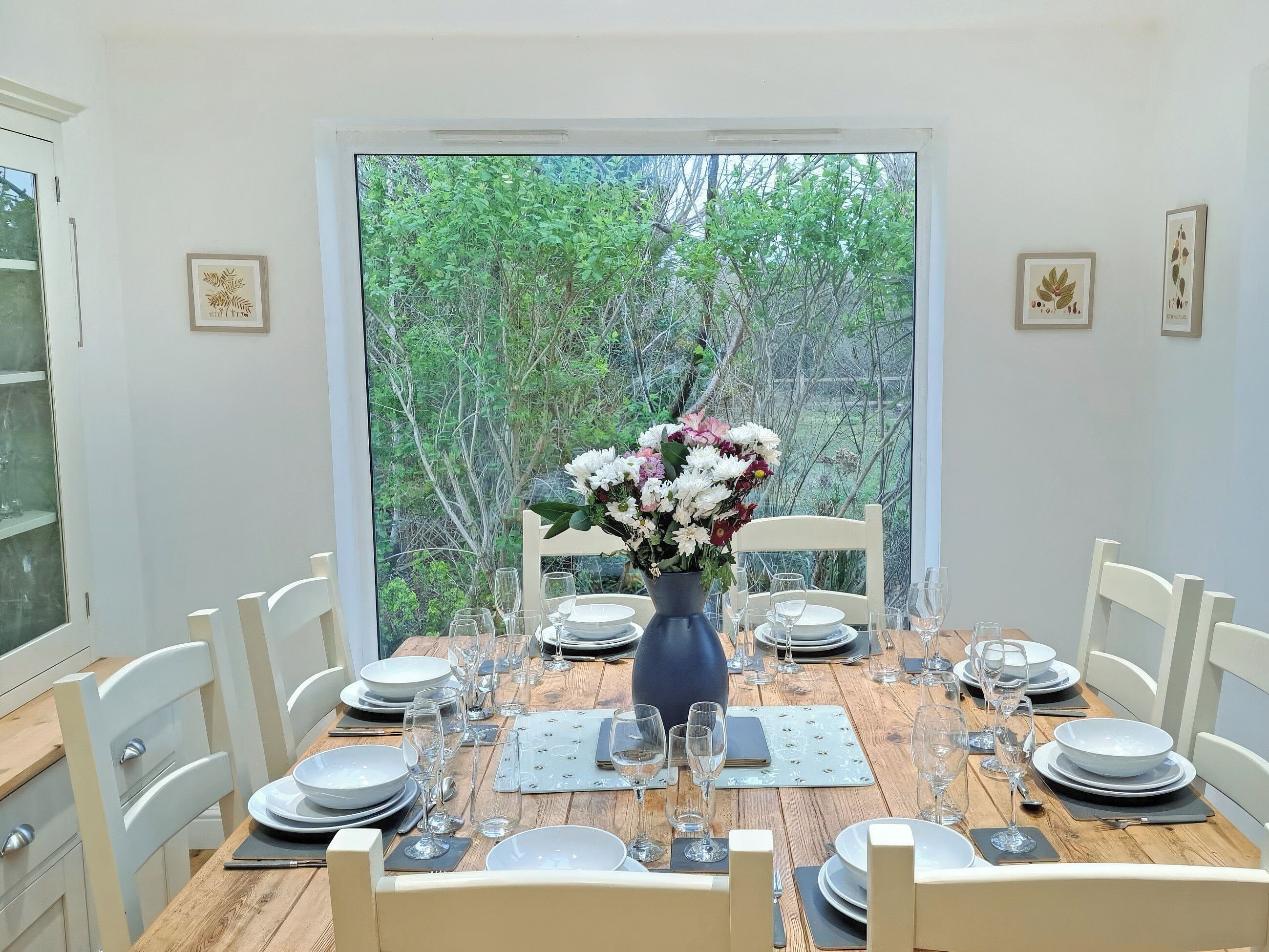 Mesa de comedor preparada para seis personas, con un florero, sillas blancas y vistas a zonas verdes a través de un ventanal grande.