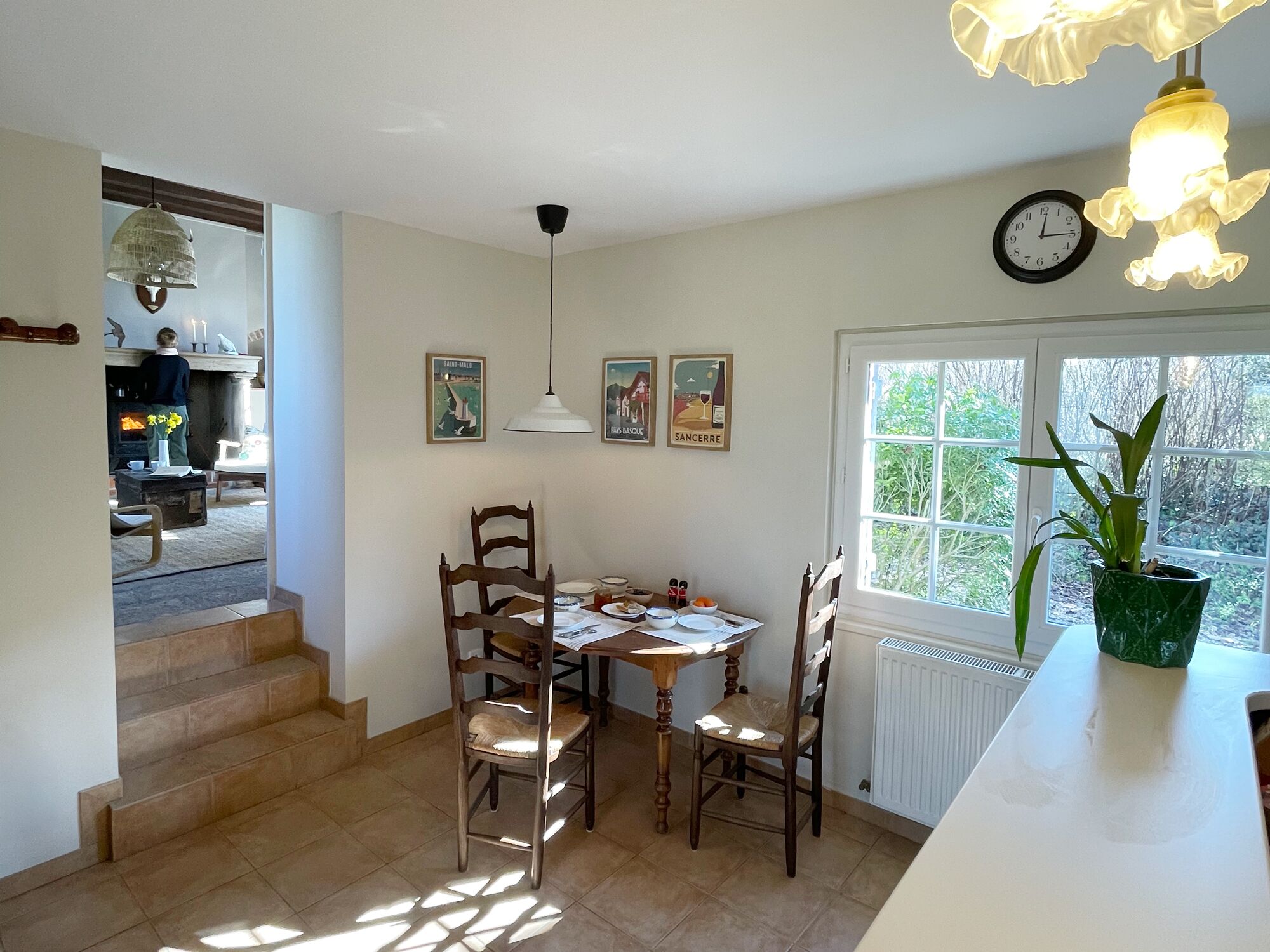 A dining area with a table, chairs, a clock, and a window with a view of greenery.