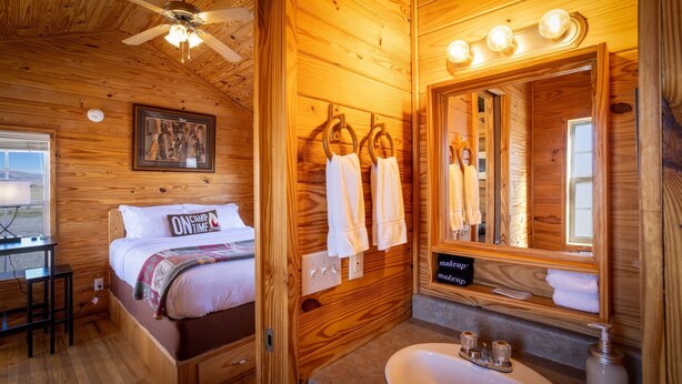 Una habitación tipo cabaña de madera con cama, escritorio, baño con lavamanos y espejo, y ventilador de techo.