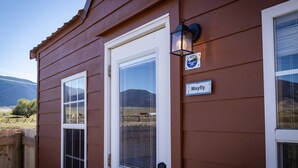 A wooden cabin with a white door, a window with a view of the mountains, and a sign that reads "Mayfly." - Mayfly Cabin (Cabin #3) (Park County)