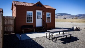 A small wooden cabin with a front door, two windows, a bench, and a picnic table.