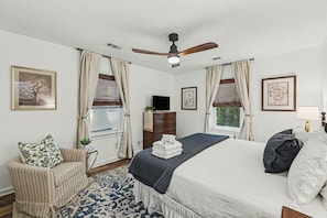 Une chambre à coucher comprenant un lit, une chaise, une commode, un ventilateur de plafond et une fenêtre avec des rideaux.