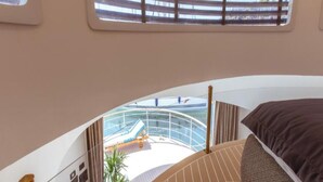 Et moderne interiør med en buet seng, udsigt til poolområdet og et vindue med vandrette persienner.