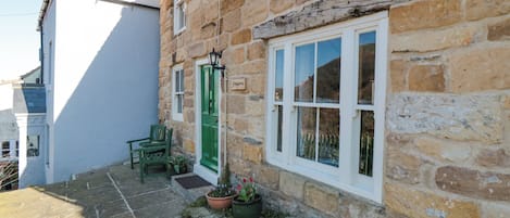 A stone building with a green door and a white window. There is a chair and a potted plant outside.