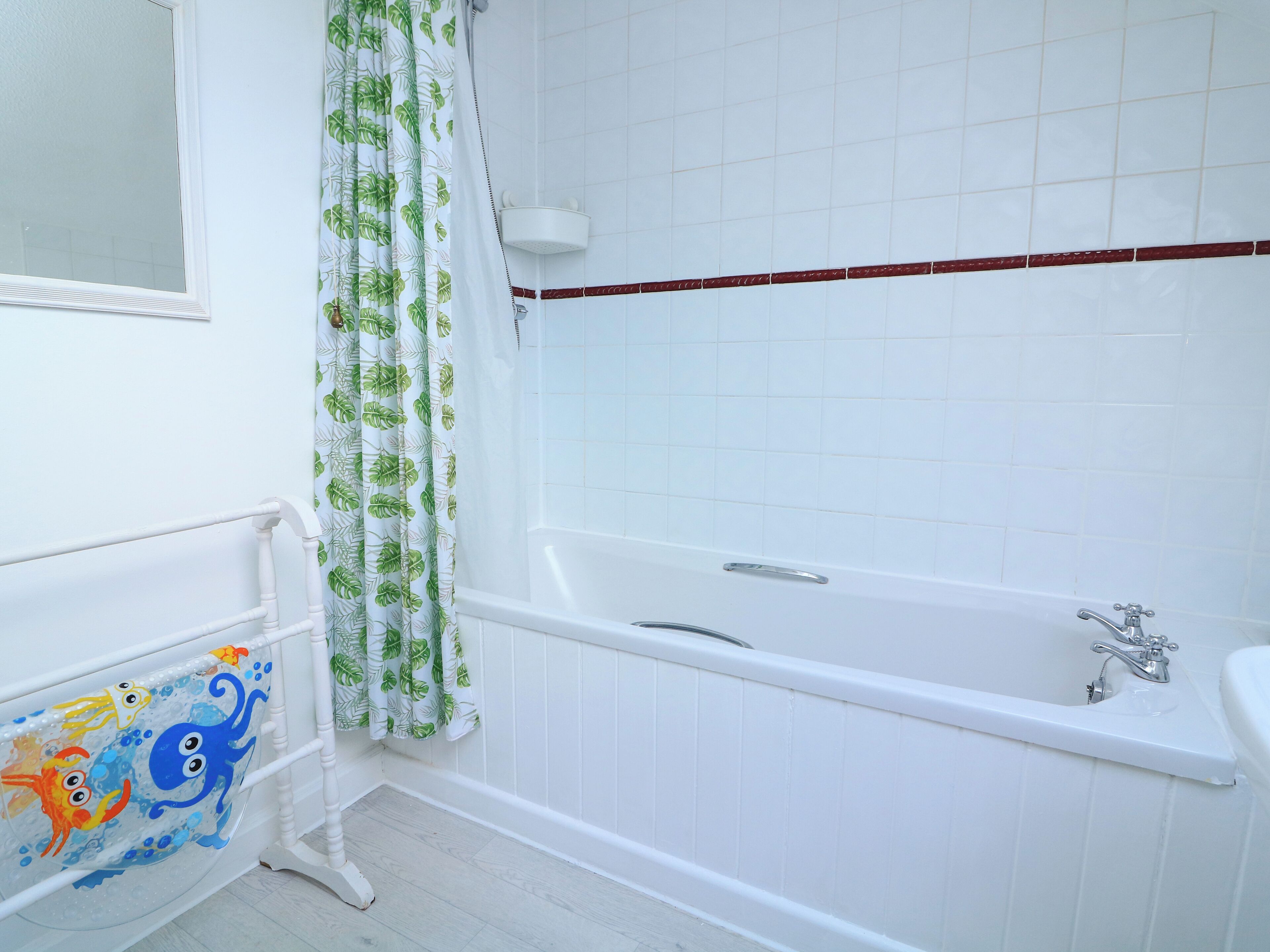 Un baño con una bañera blanca, una cortina de ducha con estampado de hojas y una pared de azulejos blancos.