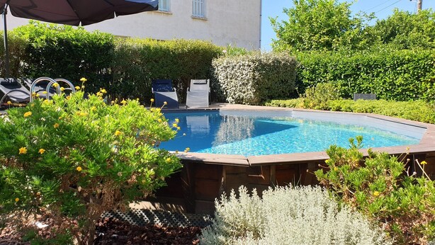 A swimming pool surrounded by lush greenery and a building in the background. - A l'ombre du clocher - Gîte with heated pool (Calenzana)