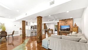 A spacious living area with a large sofa, wooden pillars, a kitchen in the background, and a TV mounted on the wall. - Private Fairhope Waterfront Retreat, Two Complete Residences, Sleeps Up to 14 (Fairhope)