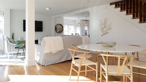 A modern living room with a round dining table, striped sofa, wooden staircase, and a large mirror on the wall.