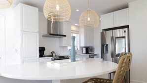 A modern kitchen with white cabinetry, a central island, stainless steel appliances, wicker chairs, and pendant lighting.