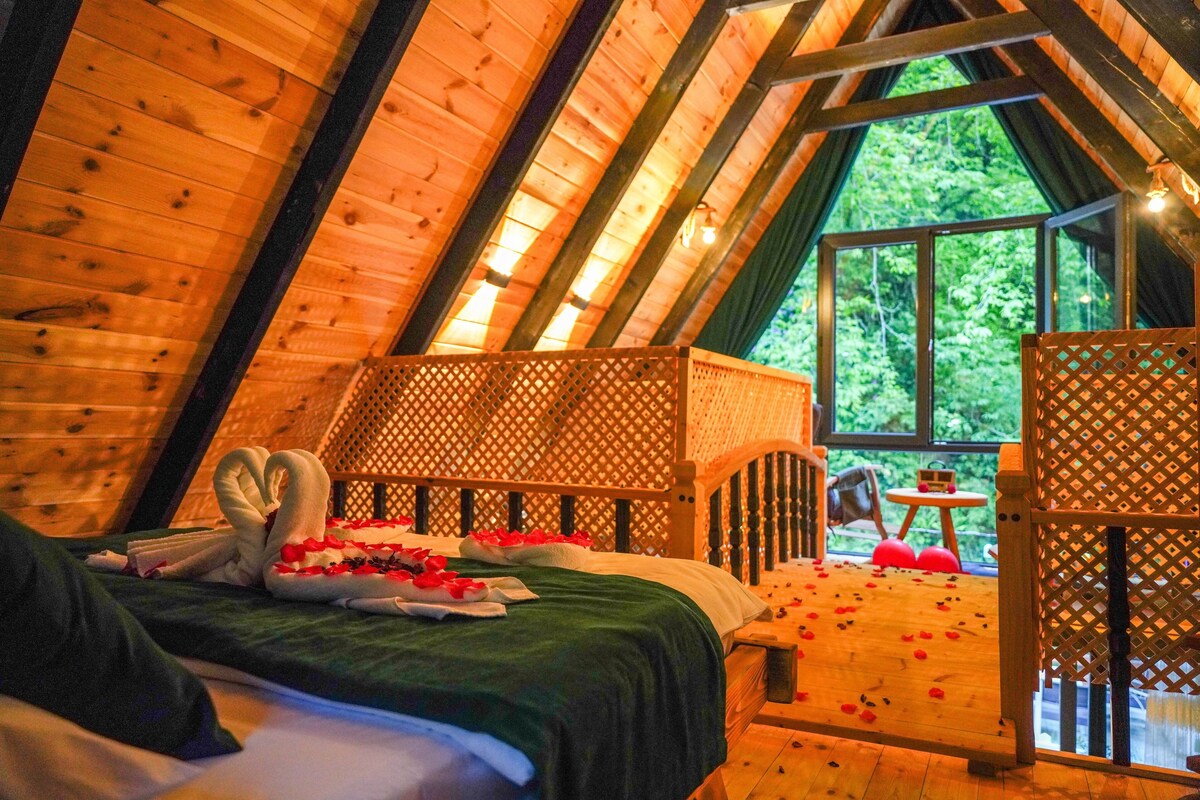 Een knus slaapkamer in een houten hut met een bed, een klein tafeltje en een raam met uitzicht op groen.