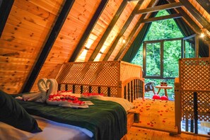 Een knus slaapkamer in een houten hut met een bed, een klein tafeltje en een raam met uitzicht op groen.