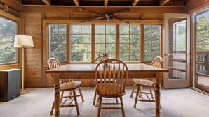 Une salle à manger en bois avec une grande table et quatre chaises, de grandes fenêtres donnant sur la forêt et un ventilateur de plafond.