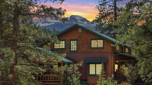 Une cabane en rondins à deux étages, surmontée d’un toit végétalisé, entourée d’une forêt dense et avec des montagnes en arrière-plan.