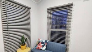 Une petite pièce avec un canapé bleu, une petite table d’appoint ronde et jaune sur laquelle est posée une plante en pot, et deux fenêtres équipées de stores.