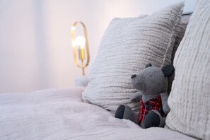 A bed with a grey teddy bear wearing a plaid shirt, white pillows, and a bedside lamp with a gold base.
