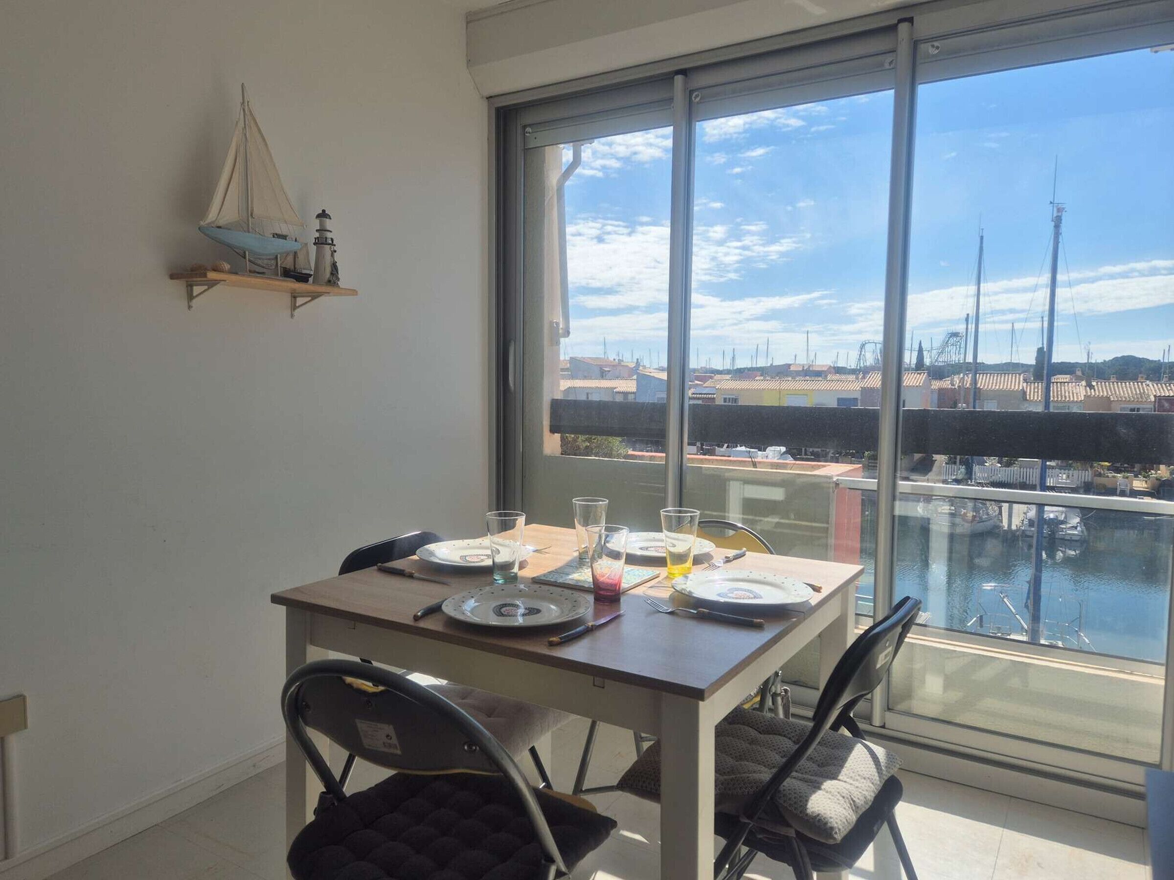 A dining area with a table set for two, a view of a marina through large windows, and a small sailboat decoration on the wall.