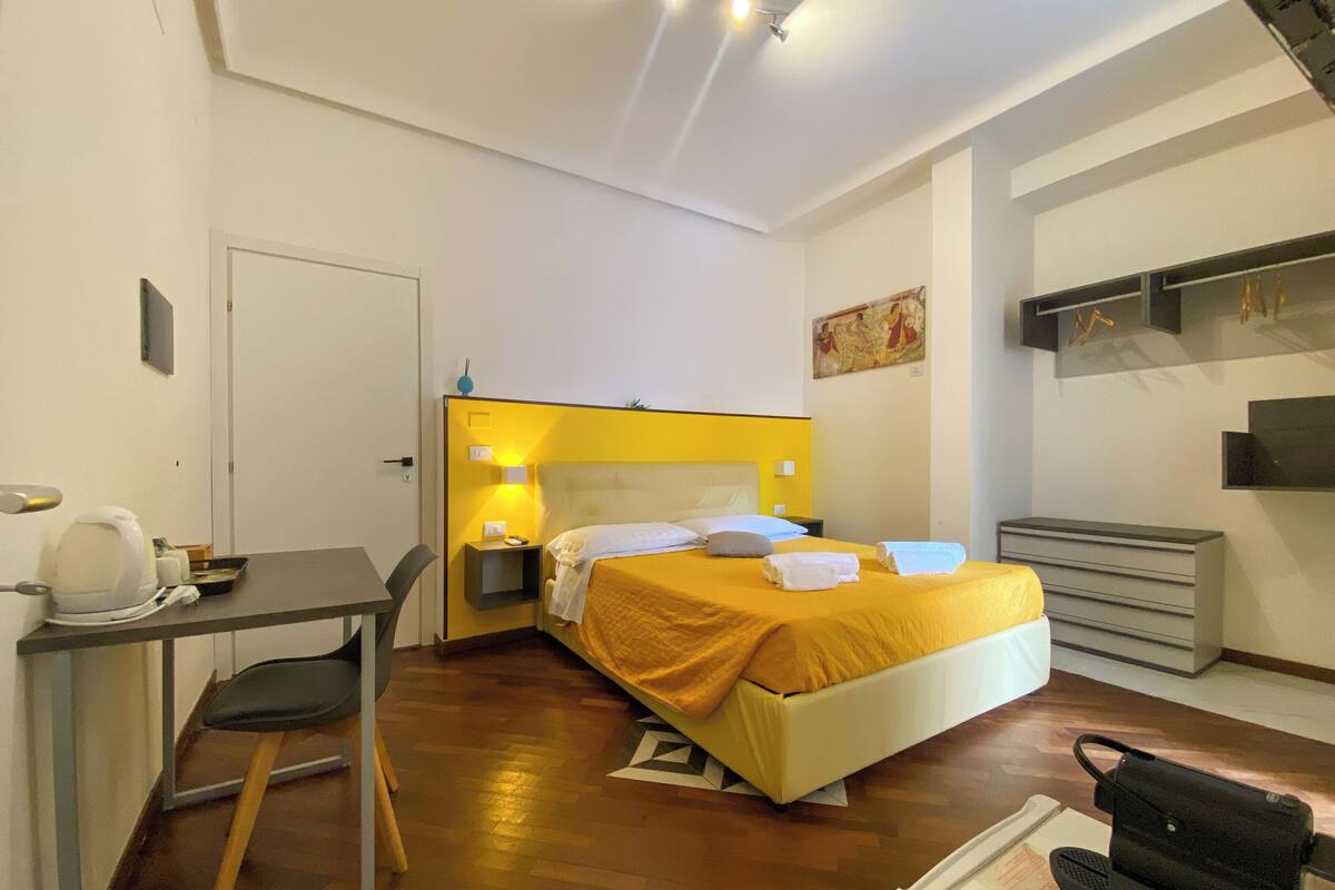 A hotel room with a bed, bedside table, chair, and a small desk with a kettle.