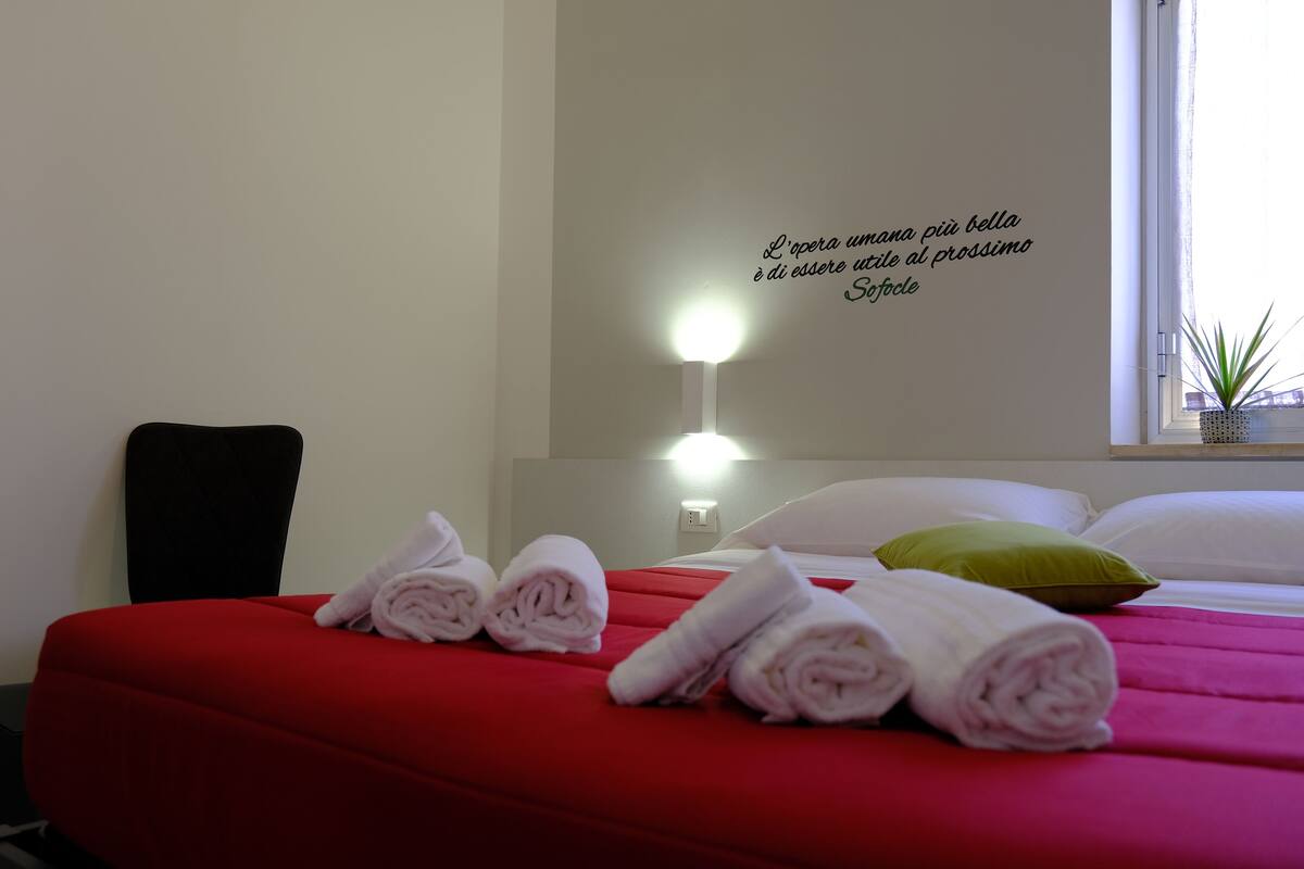 A hotel room with a red bed, white pillows, and a wall quote in Italian.