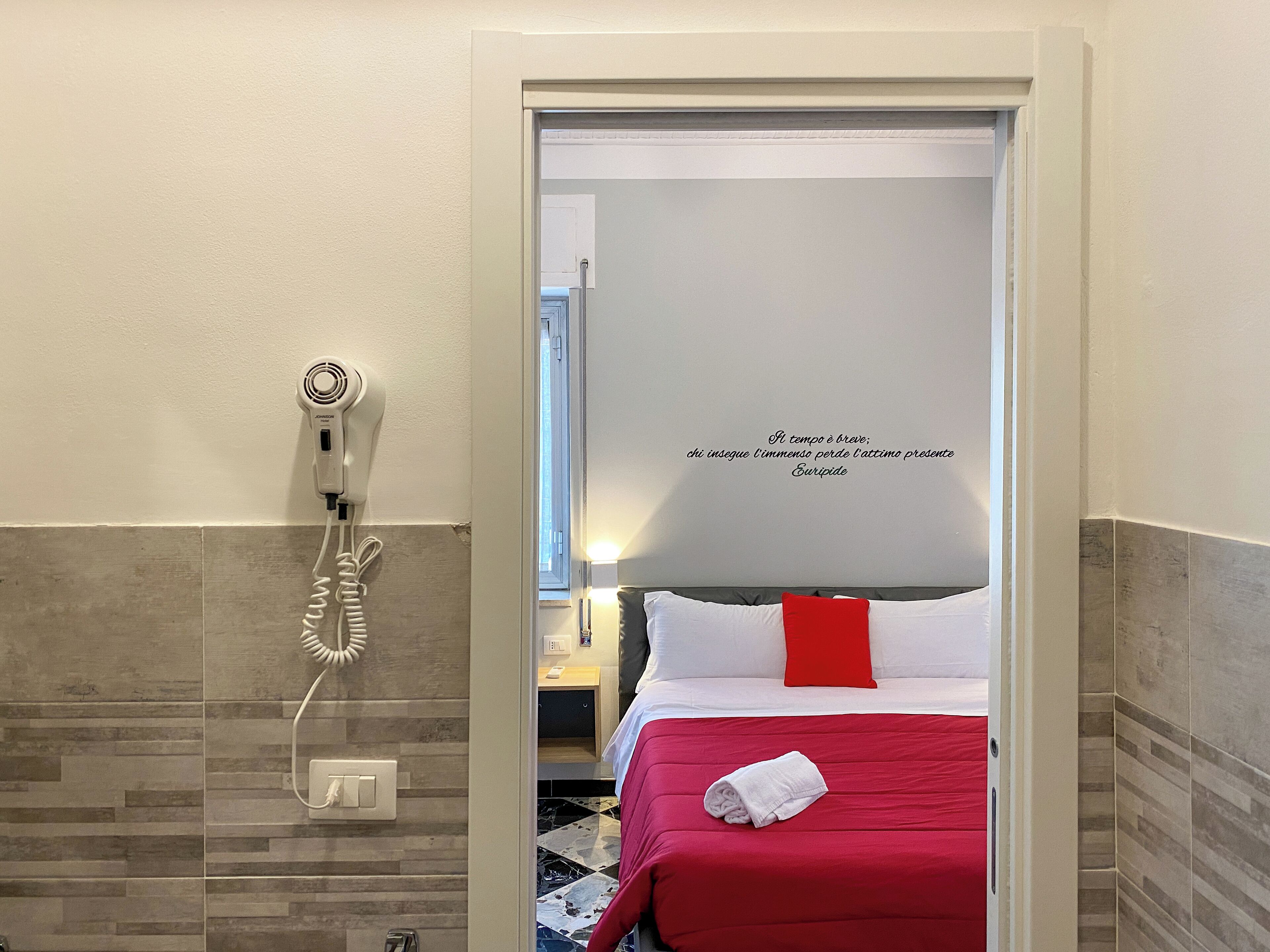 Una habitación de hotel con cama, almohada roja y blanca, toalla blanca y secador de pelo en la pared.