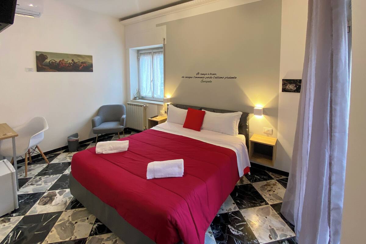 A hotel room with a bed, a chair, a desk, a window, and a painting on the wall.