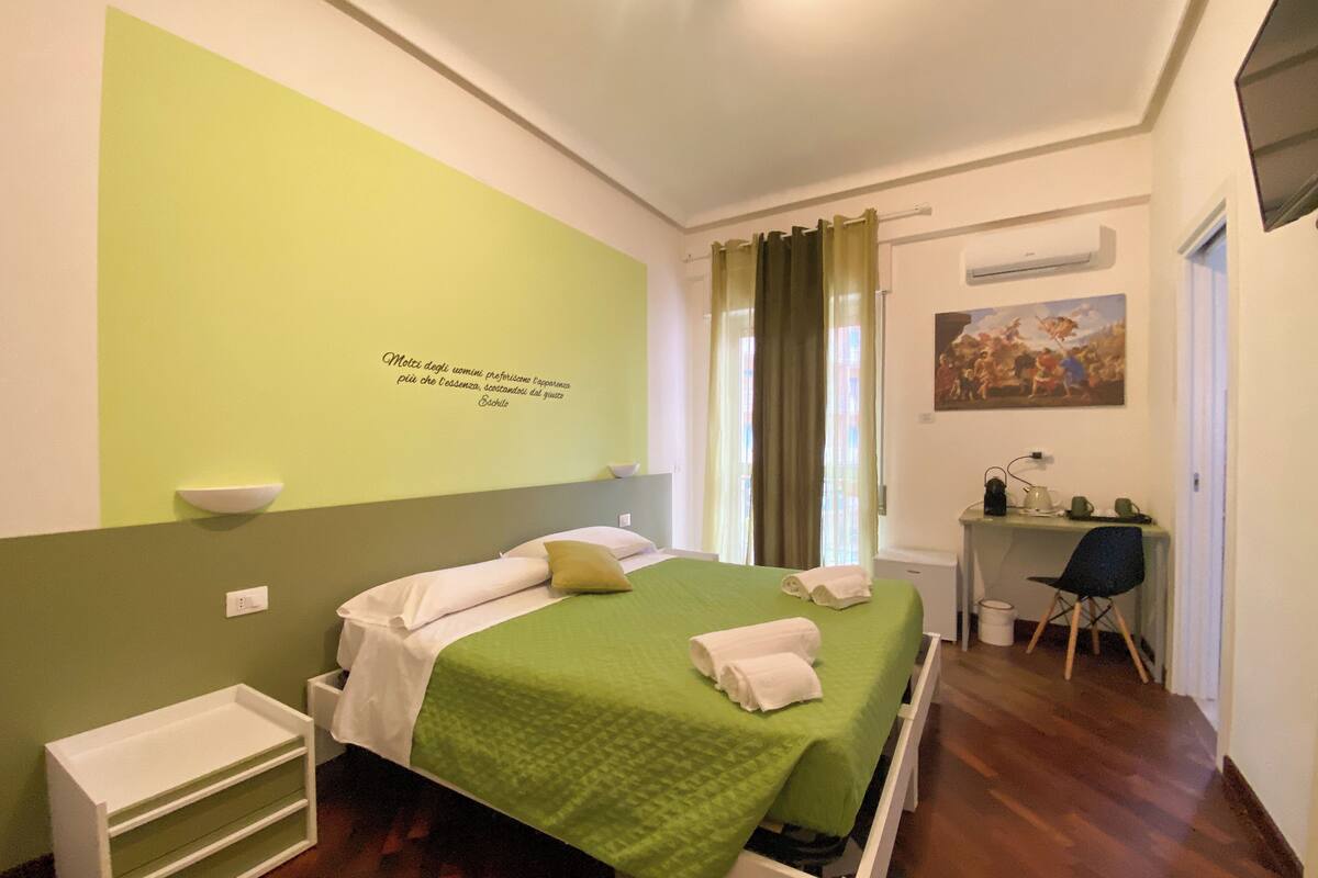 A hotel room with a bed, a desk, a chair, a small shelf, and a painting on the wall.