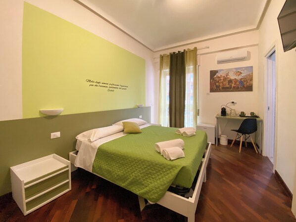 A hotel room with a bed, a desk, a chair, a small shelf, and a painting on the wall. - Al Teatro Greco (Syracuse)