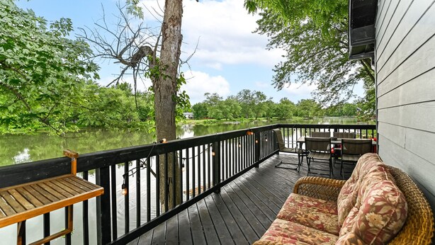 A wooden deck with a wicker sofa and a small table, overlooking a calm lake and surrounded by trees. - Riverfront Hideaway/Kayaks/Pet‑Friendly/Beautiful Views! (Noblesville)