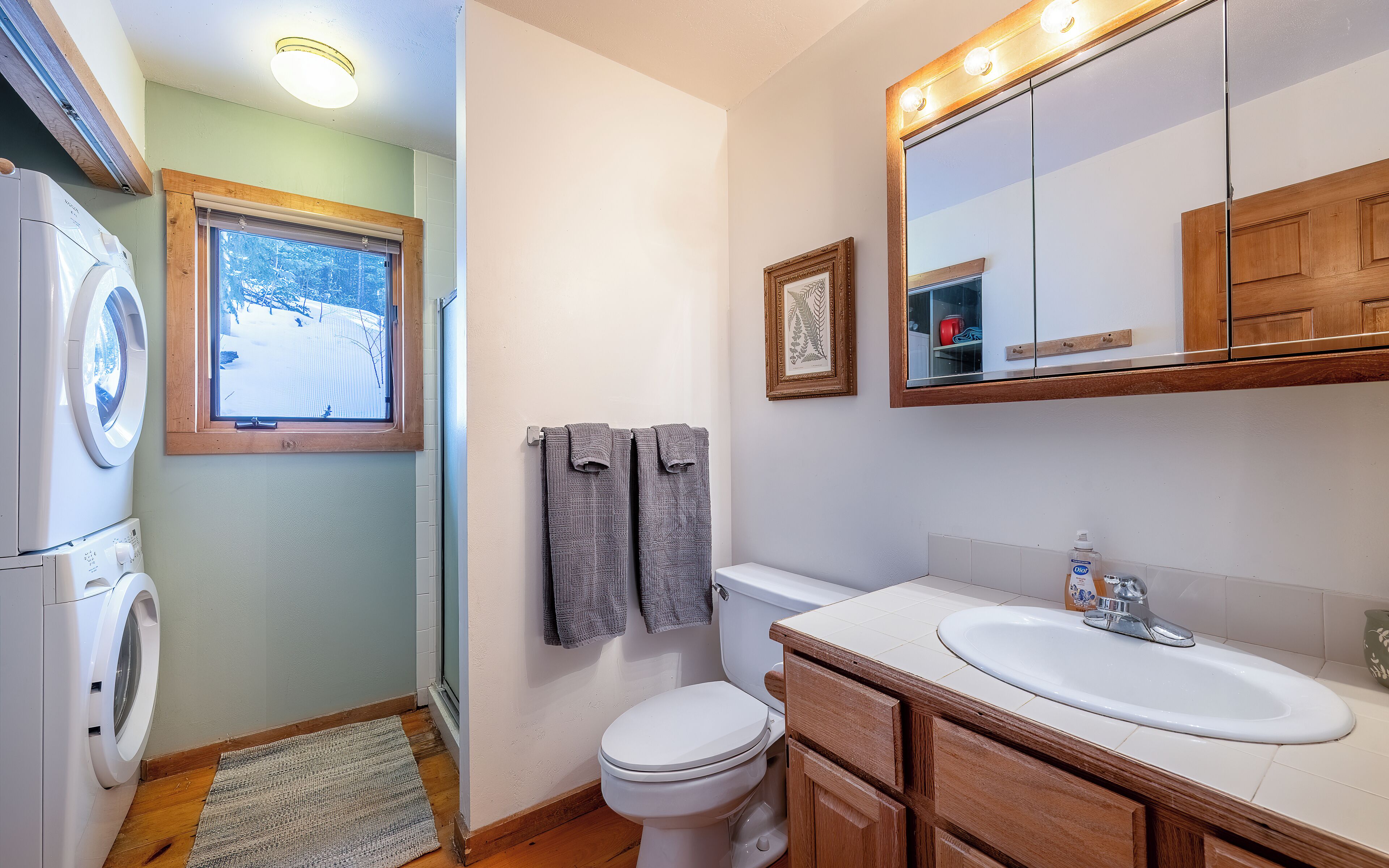 A bathroom with a washing machine, a toilet, a sink, and a window with a view of a snowy landscape.