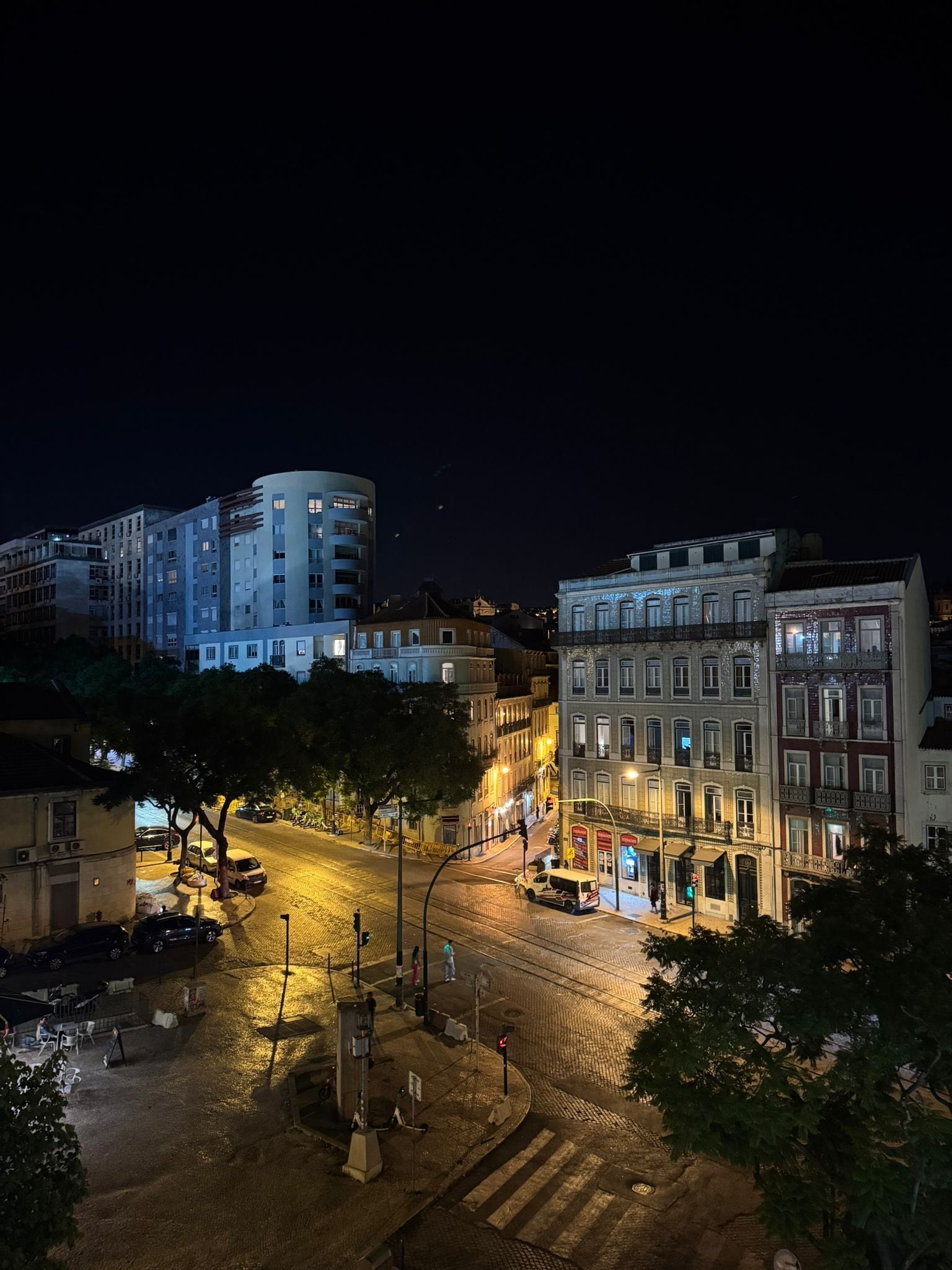 A nighttime cityscape with illuminated buildings, a cobblestone street, and a pedestrian crossing.