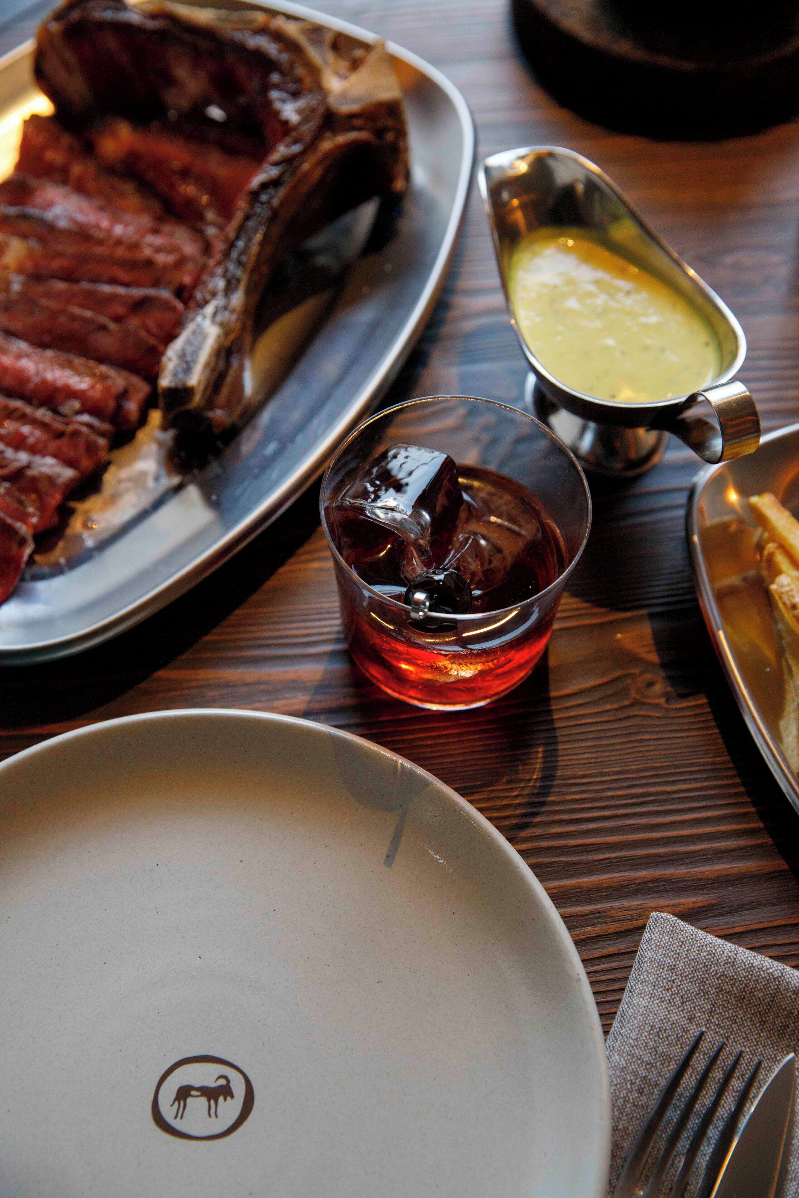 A plate of sliced meat, a glass of red beverage, and a small dish of sauce on a wooden table.