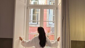 Eine Person in einem weißen Gewand steht oben an einer engen Treppe mit Blick auf ein Gebäude außerhalb.