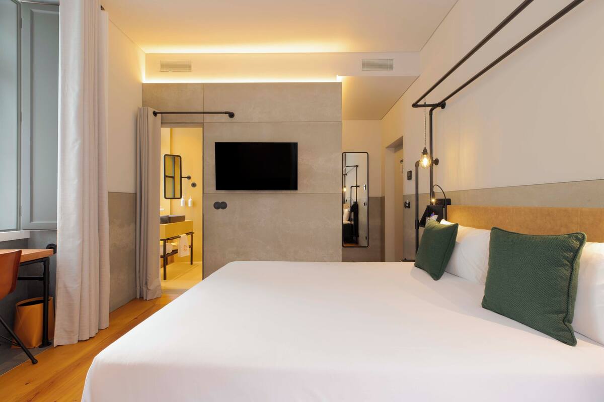 Habitación de hotel con cama, escritorio, silla, televisor, espejo y baño visible a través de una puerta abierta.