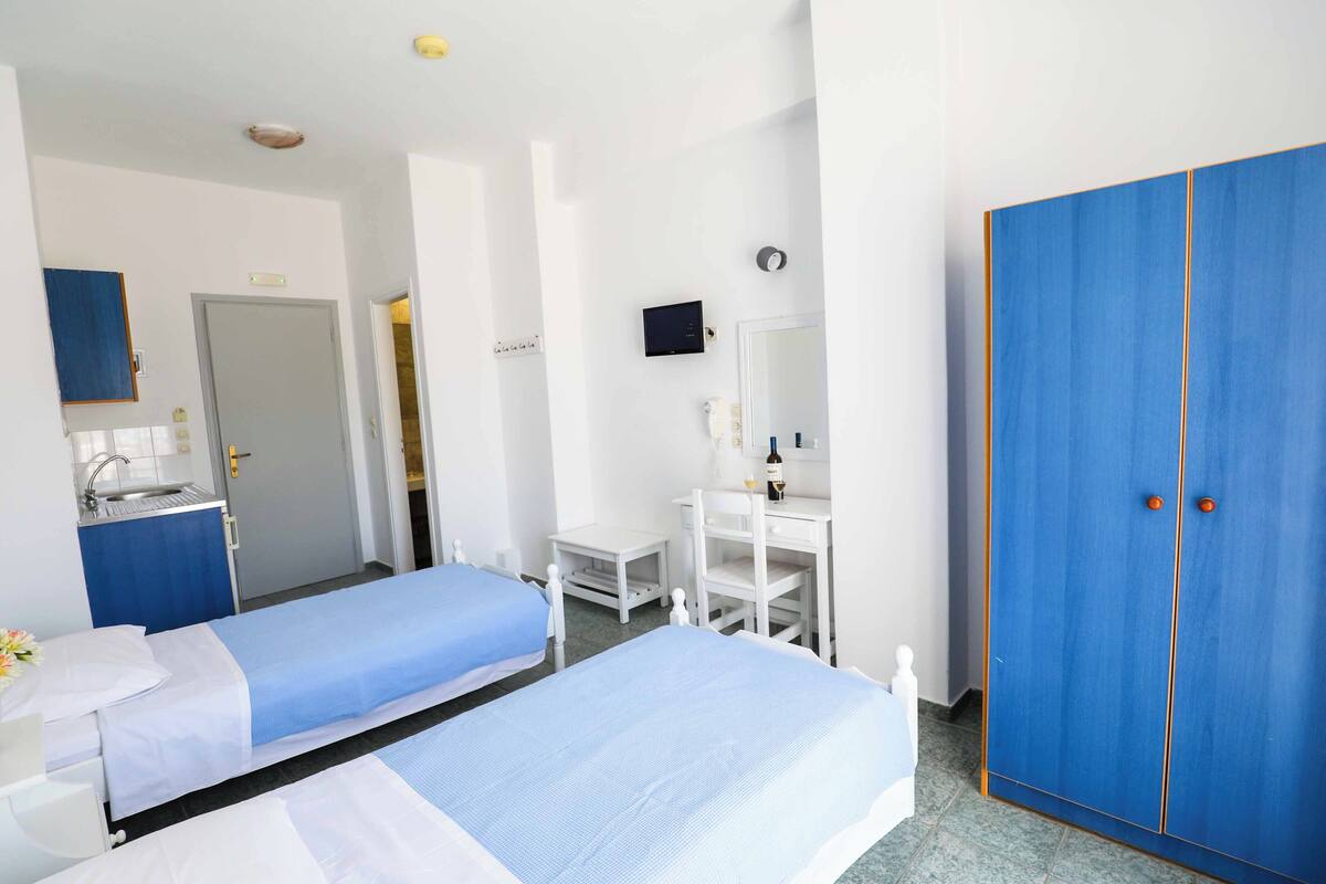 A hotel room with two single beds, a small desk, a mirror, a wardrobe, and a bathroom with a sink and toilet.