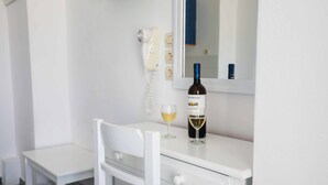 A white desk with a bottle of wine and a glass, a white chair, a white telephone, and a mirror on the wall. - Finikas Santorini (Kamari)