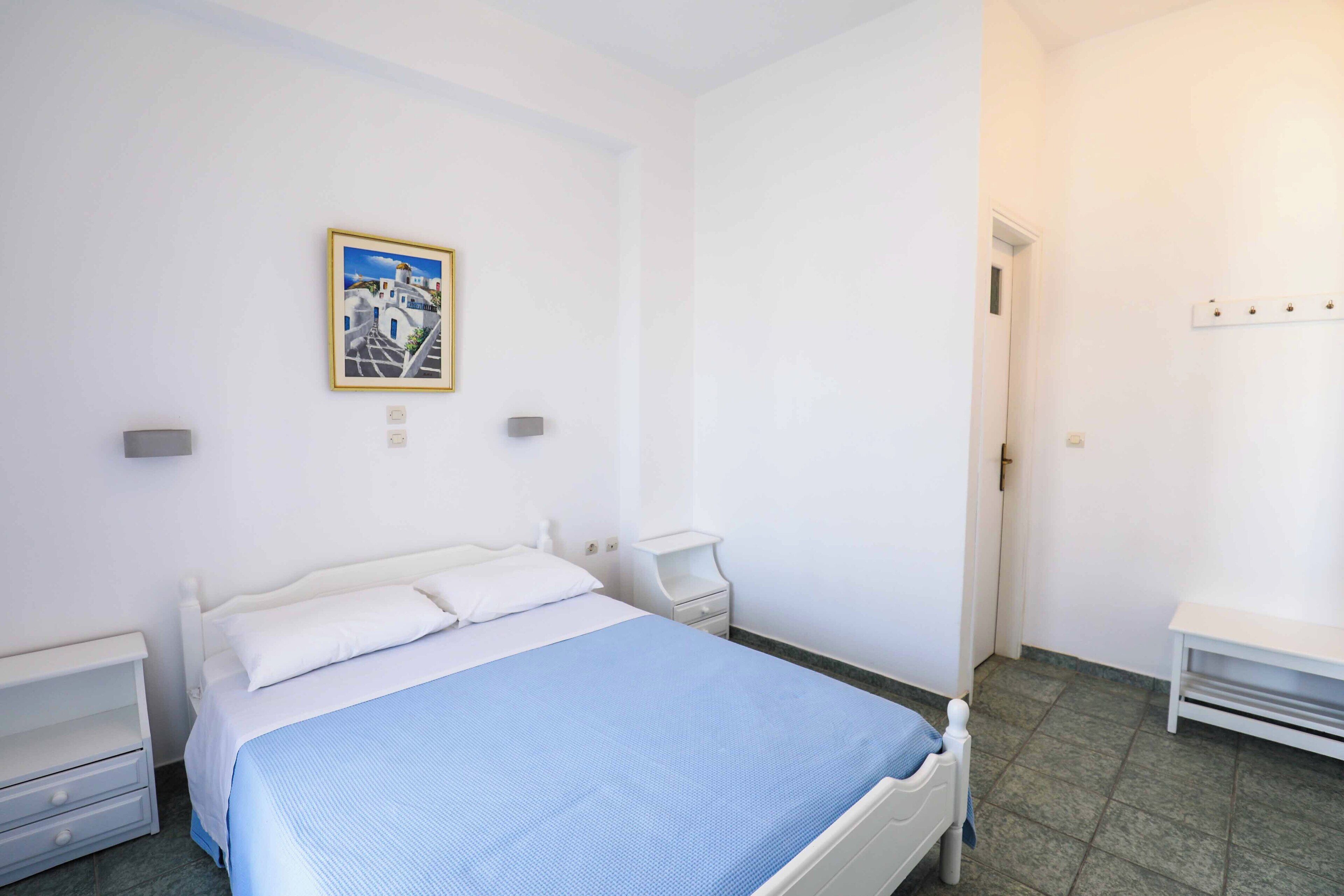 A bedroom with a bed, bedside tables, a nightstand, a wall-mounted shelf, and a door.