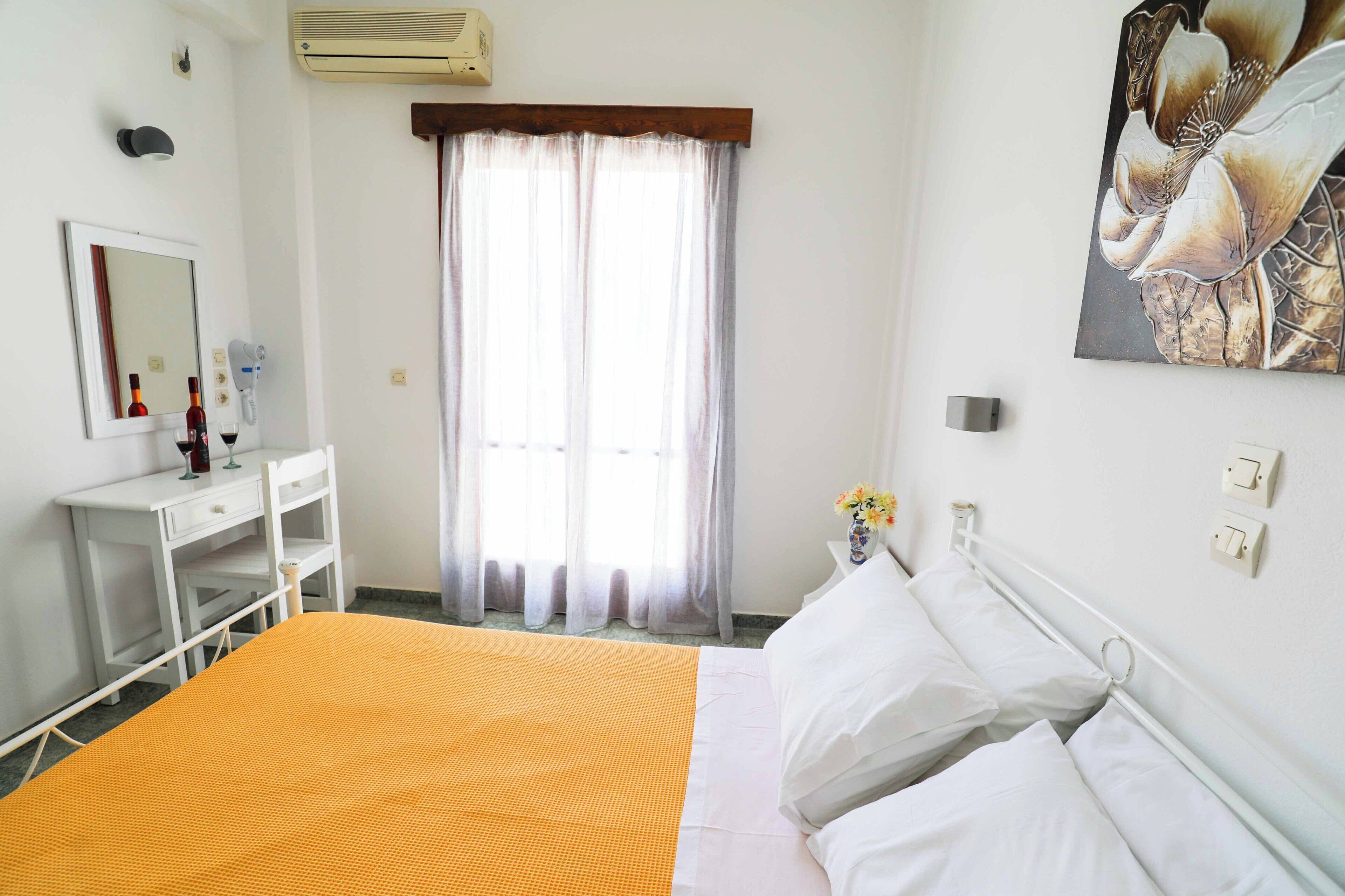 A hotel room with a bed, a desk, a mirror, a window with curtains, and a wall-mounted air conditioner.