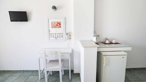 A small, white room with a desk, chair, and a refrigerator. There is a flat-screen TV mounted on the wall and a framed picture above the desk. - Finikas Santorini (Kamari)