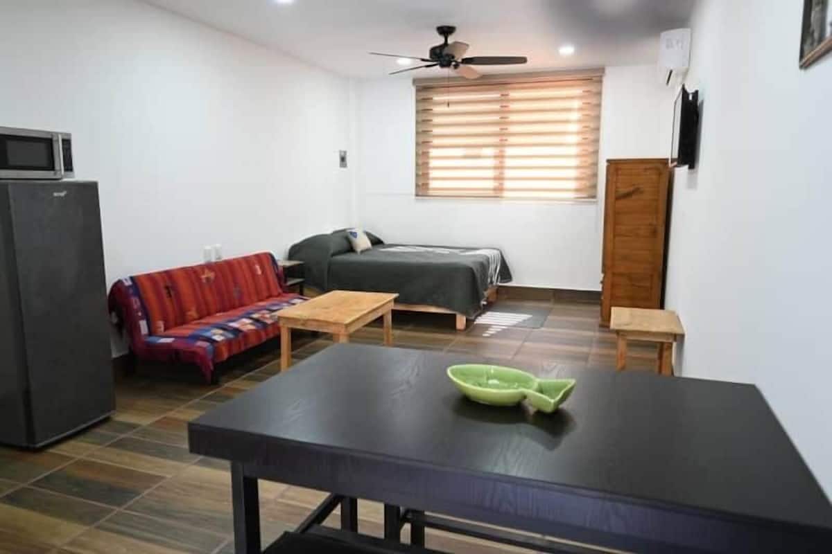 A compact room with a bed, a sofa, a dining table, a ceiling fan, and a window with blinds.
