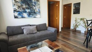 Ein Wohnzimmer mit grauem Sofa, einem Glastisch mit Papieren, einem großen abstrakten Gemälde und einer Holztür.