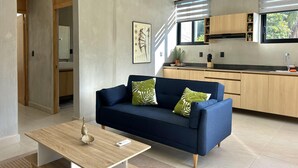 Una sala de estar moderna con un sofá azul oscuro, una mesa de centro de madera y una zona de cocina con armarios de madera y una ventana con persianas.