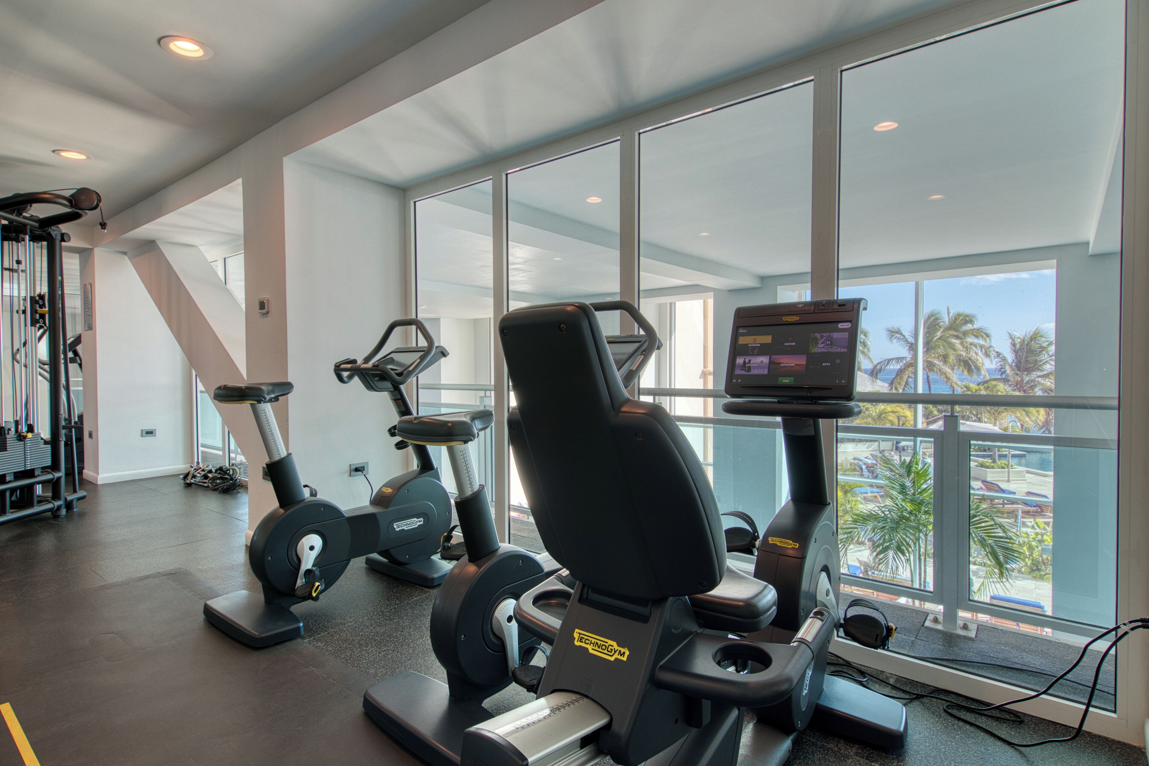 A gym with cardio machines, weight equipment, and large windows offering a view of palm trees and a parking lot.