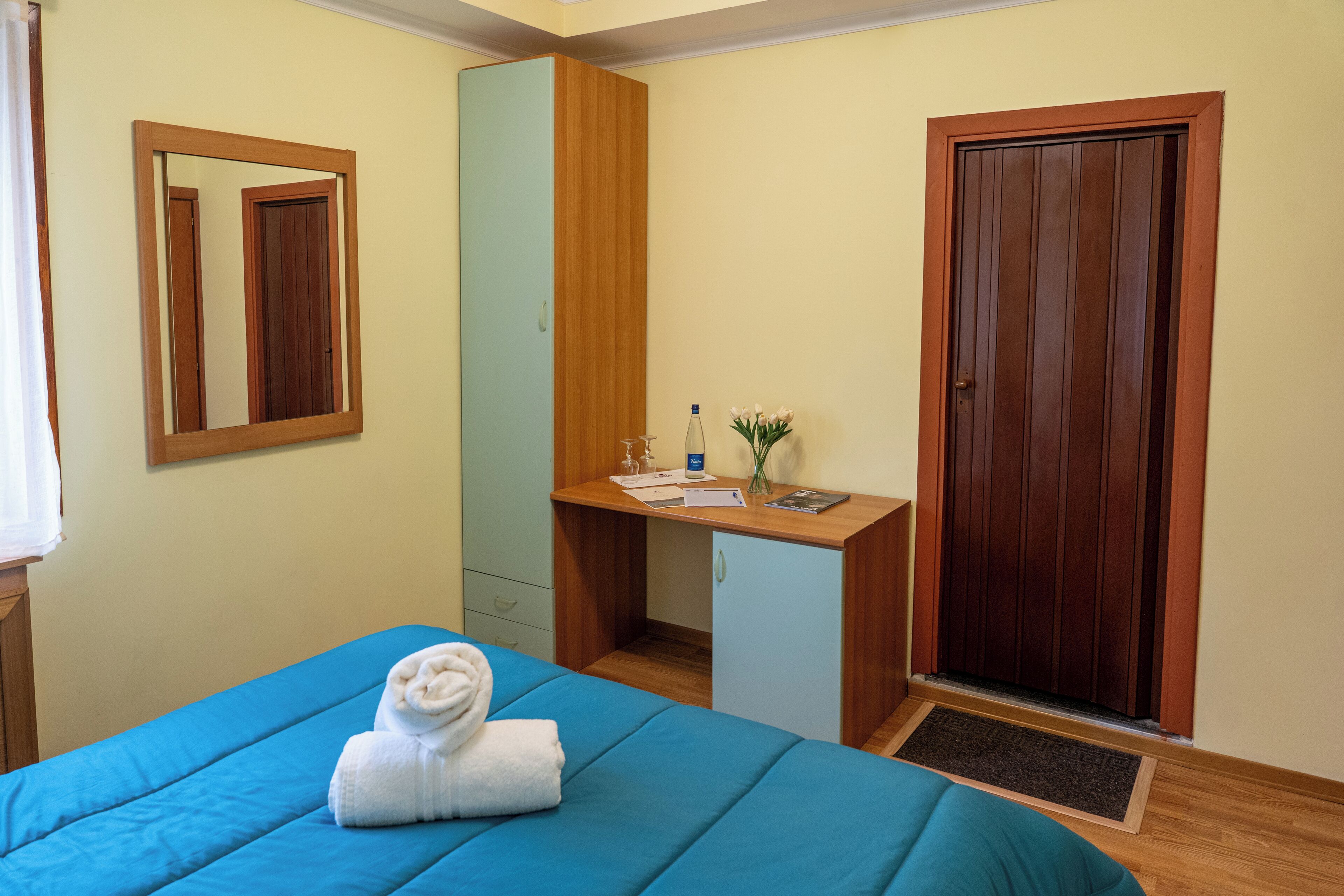 A hotel room with a bed, a desk, a mirror, a wardrobe, and a door.