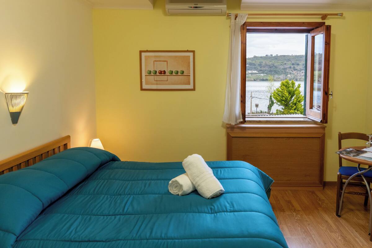 A bedroom with a bed, a window with a view, a chair, and a wall-mounted lamp.