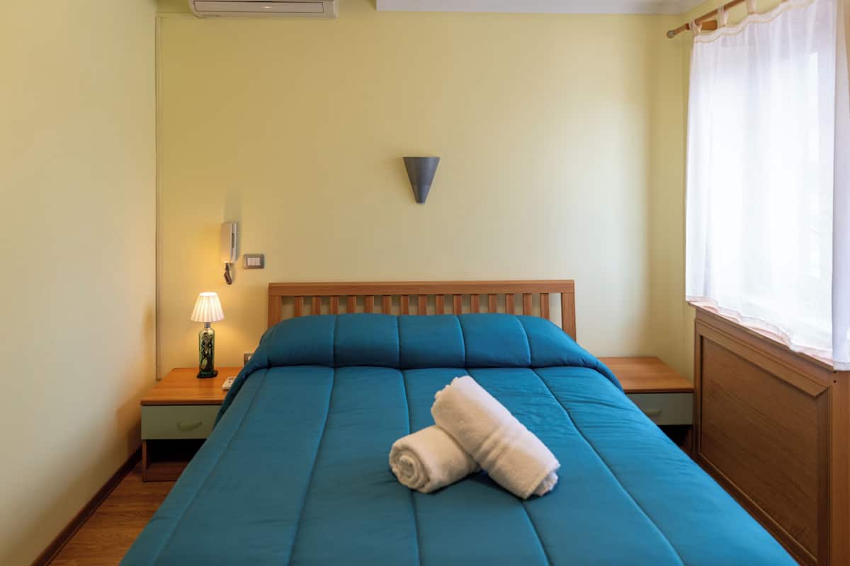 A neatly made bed with a blue mattress and a rolled white towel, a bedside lamp, a small wooden nightstand, and a wall-mounted light fixture.