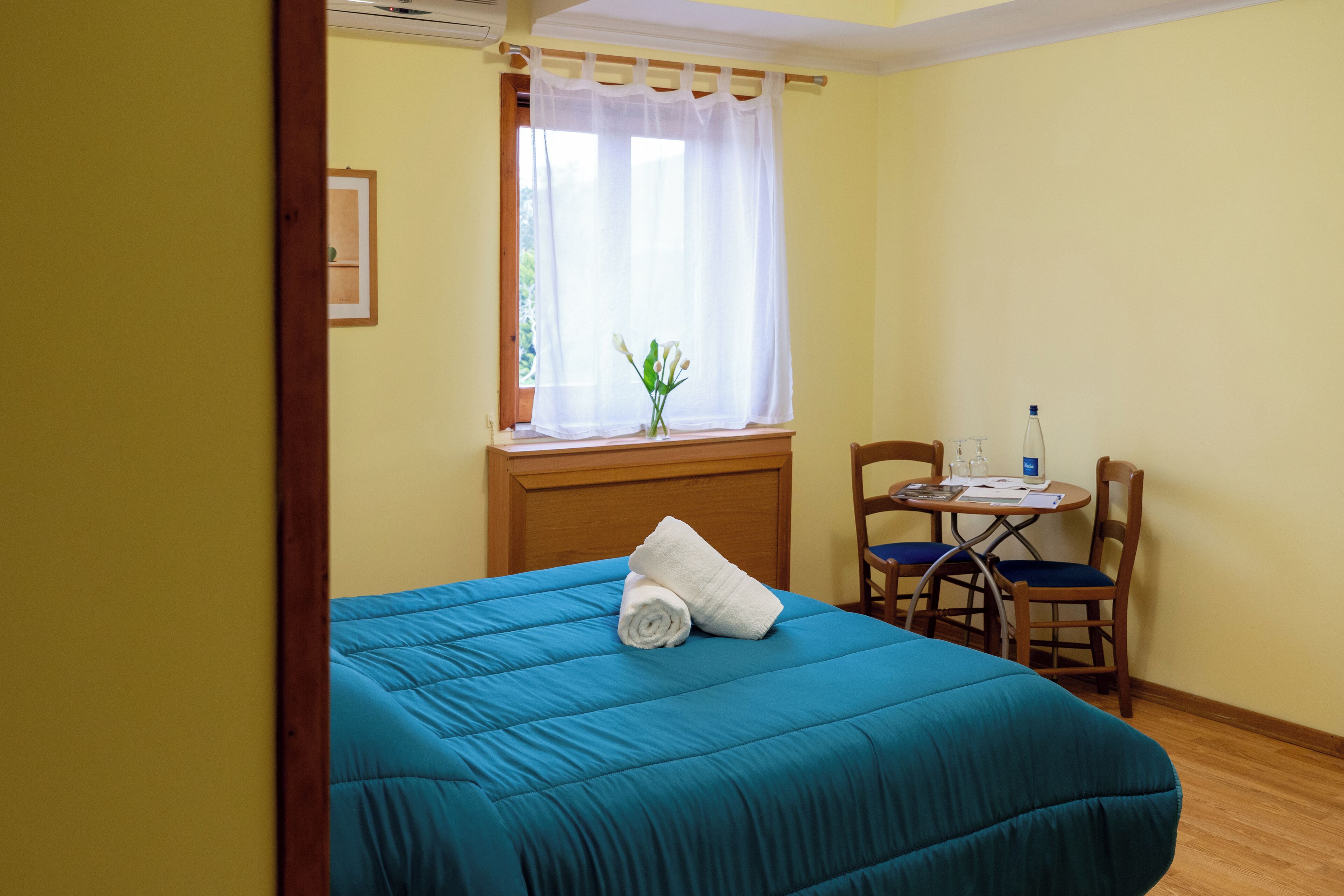 A room with a bed, a small table with chairs, a window with a curtain, and a plant on the windowsill.