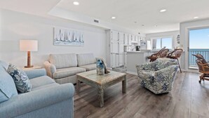 A modern living room with a sofa, armchair, and coffee table, a kitchen area, and a balcony with wicker furniture. - Corner, Oceanfront Balcony- Pool- Upscale Design (North Myrtle Beach)
