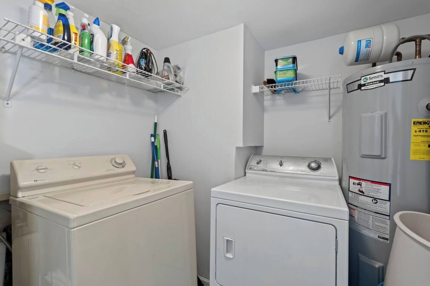 A laundry room with two washing machines, a shelf with cleaning supplies, and a water heater.