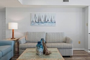 Un salón con un sofá a rayas, una mesa de centro con objetos decorativos, una lámpara y un cuadro de barcos de vela en la pared.
