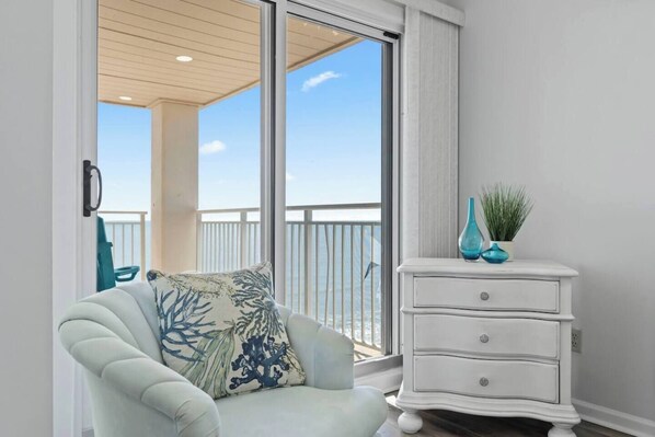 A room with a view of the sea, a white dresser with decorative items, and a light-colored armchair.