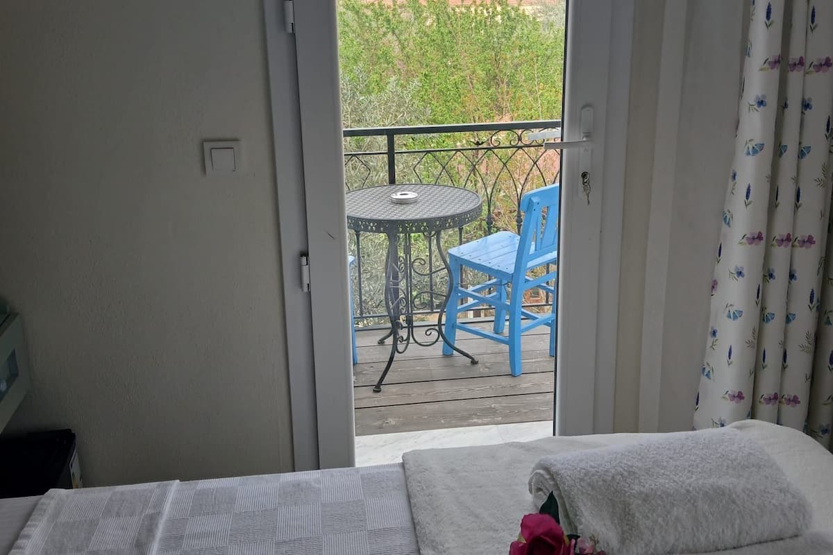 A hotel room with a bed, a small table with chairs, and a view of a balcony with a table and chairs outside.